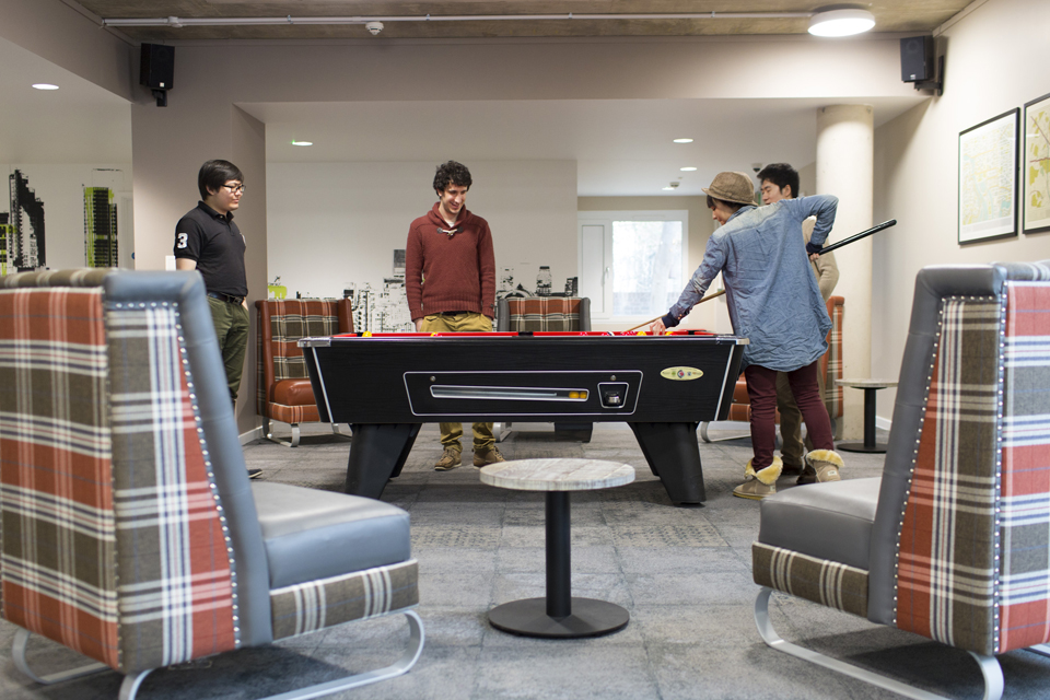Two comfy chairs in the foreground, with a group of students playing a game of pool in the background.